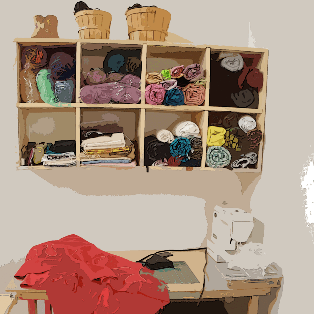 system, filled with rolls of fabric. Atop are three baskets, each one larger than the one to the left. Below the cubby is the top of a desk, on which sits a white sewing machine on the right. The eye follows a wire from the back of the sewing machine to the pedal, also sitting on the desk, on top of a green cutting mat. To the immediate left lies a large red swath of fabric, it dangles over the front and left edges of the table.